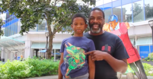 One older Black male wearing a blue tee shirt and his young son wearing a blue tee shirt with a lizard, standing in front of a sculpture talking about Sculptures and Stories.