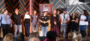 9 Adults standing in a line on stage reviewing their stories