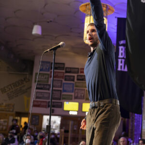 John Valentyn holds up his left arm to the ceiling. He is speaking into the microphone. He is wearing a blue long sleeve tucked into brown pants.
