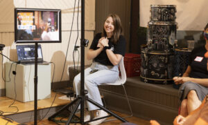 Lauren, a young white woman is sitting in a chair with her hands clasped in front of her smiling at the camera.