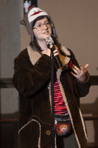 Storyteller wearing a hat with a monkey on it, glasses, and brown jacket holding microphone.