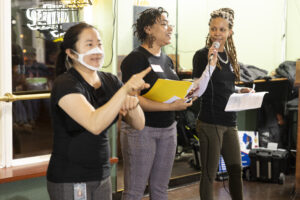 Alea McHatten, Kim Shine, and ASL interpreter stand in a row. Kim speaks into a microphone.