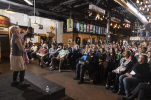 Storyteller on stage holding left hand out while speaking into the microphone in front of of crowded room.