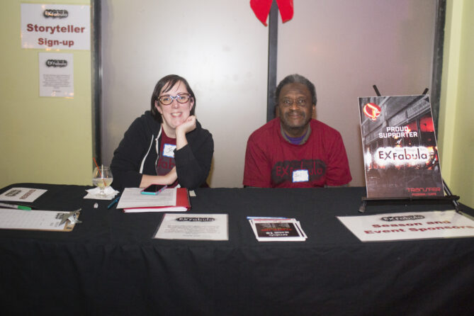 Jess Betts sitting on the right next to another volunteer. Jess has brown hair, glasses and is wearing a black hoodie.