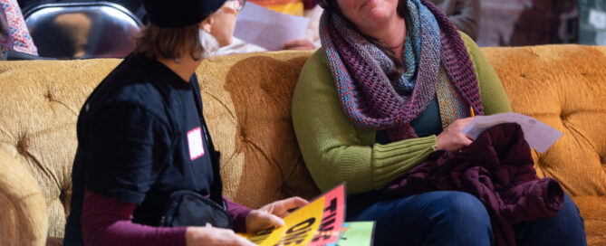 Two people sit next to each other on a yellow couch, laughing and enjoying the event.